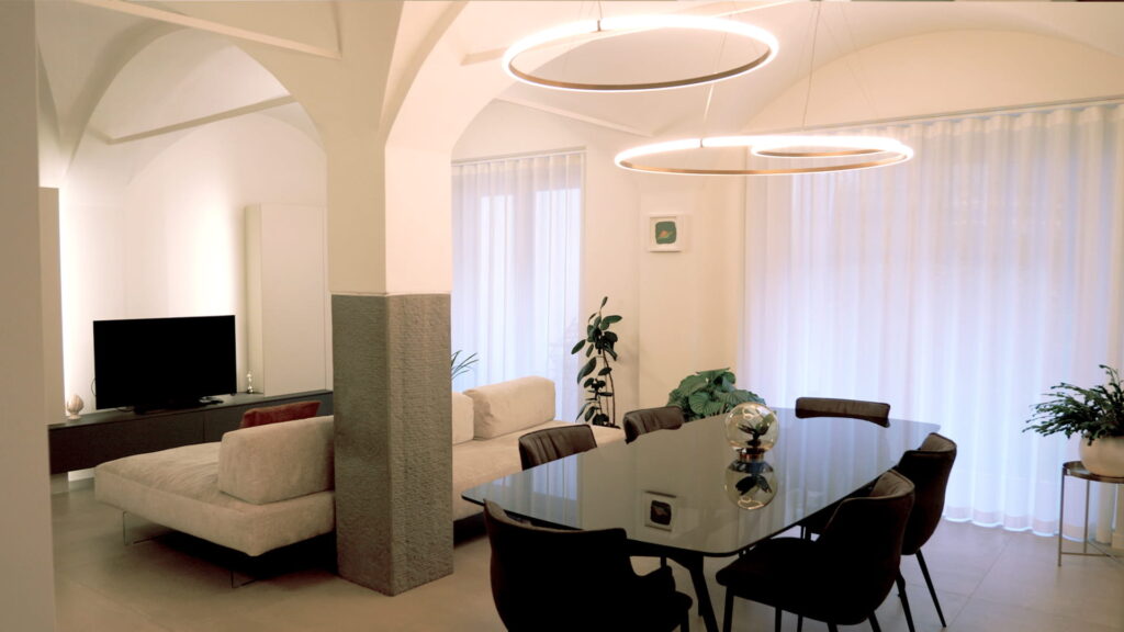 Open space residenziale con zona pranzo e soggiorno separati da colonna in pietra grezza, soffitti a volta con lampadari circolari in ottone e tende in lino bianco