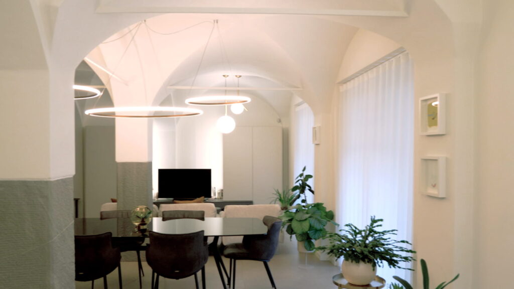 Prospettiva dell'open space con volte a crociera, tavolo da pranzo con sedie in similpelle scura, cucina a isola sullo sfondo e composizione di piante