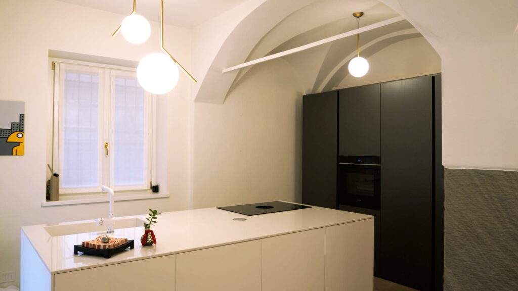 Cucina a isola bianca con piano cottura a induzione integrato, colonne attrezzate in grigio antracite e lampade a sfera su asta in ottone, soffitto a volta