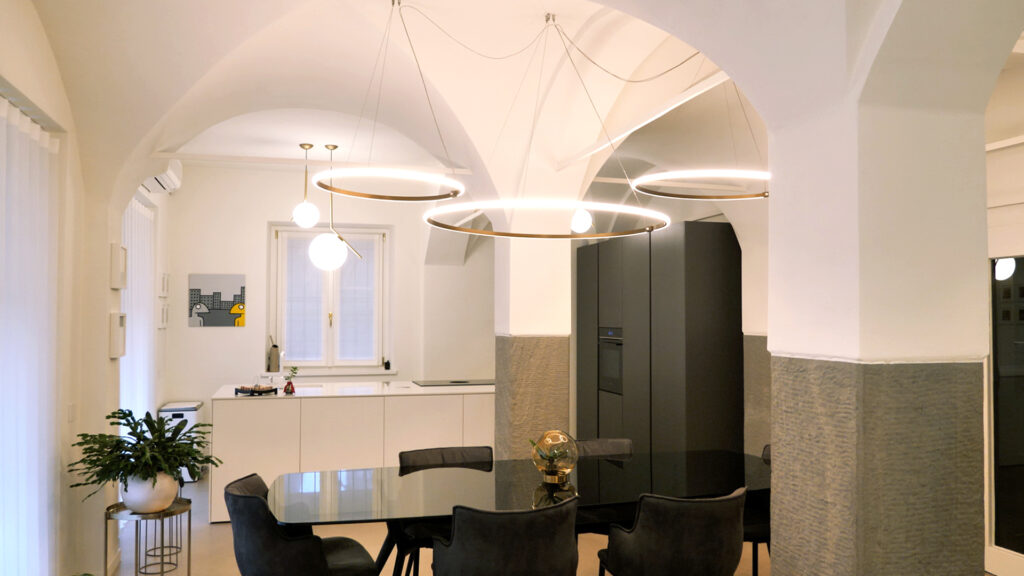 Open space con tavolo da pranzo in vetro scuro, sedie in velluto antracite, cucina a isola bianca e sistema di lampade circolari in ottone su soffitto a volta